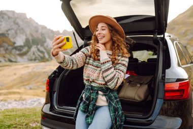 Genç gezgin kadın gün batımında arabasıyla dağlarda selfie çekiyor şık kıyafetlerini ve neşeli ruhunu sergiliyor. Yaşam tarzı, seyahat, turizm, doğa, aktif yaşam.