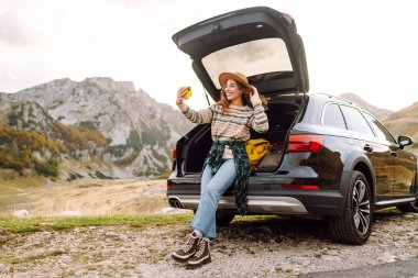 Genç gezgin kadın gün batımında arabasıyla dağlarda selfie çekiyor şık kıyafetlerini ve neşeli ruhunu sergiliyor. Yaşam tarzı, seyahat, turizm, doğa, aktif yaşam.