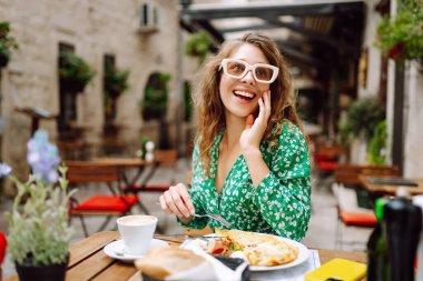 Genç bir kadın güneşli bir sabahta çiçeklerle süslenmiş büyüleyici bir kafe avlusunda kahvaltıdan zevk alır. Parmesan peyniri. Seyahat, turizm, yiyecek ve içecek..