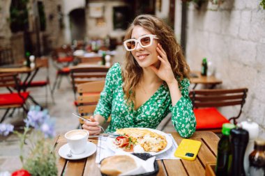 Genç bir kadın güneşli bir sabahta çiçeklerle süslenmiş büyüleyici bir kafe avlusunda kahvaltıdan zevk alır. Parmesan peyniri. Seyahat, turizm, yiyecek ve içecek..