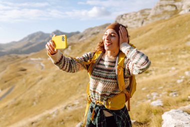 Sırt çantalı genç bir kadın mutlu bir şekilde dağlarda selfie çekiyor. Doğa özgürlüğü macerası, mutlu tatiller. Günlük tutma.