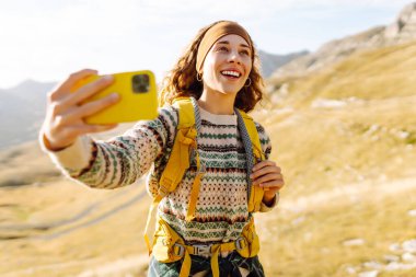 Sırt çantalı genç bir kadın mutlu bir şekilde dağlarda selfie çekiyor. Doğa özgürlüğü macerası, mutlu tatiller. Günlük tutma.