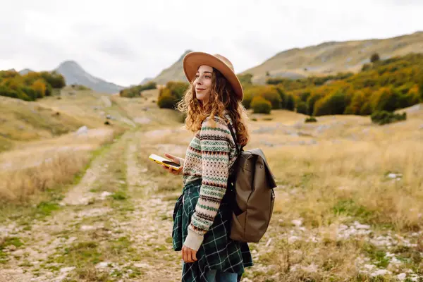 stock image A young woman is seen smiling while holding a yellow phone in an autumn landscape. She wears a cozy sweater and a wide-brimmed hat, enjoying the fresh air after hiking.