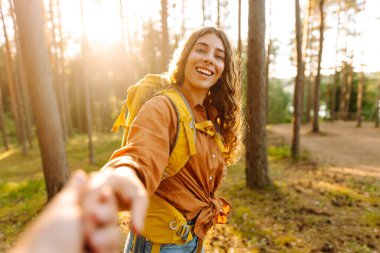 Beni takip edin! Genç bir kadın neşeli bir şekilde sırt çantasıyla güneşli orman patikalarını keşfediyor, aydınlık bir günde bir arkadaşıyla el ele tutuşuyor. Tatil, turizm. Yürüyüş.