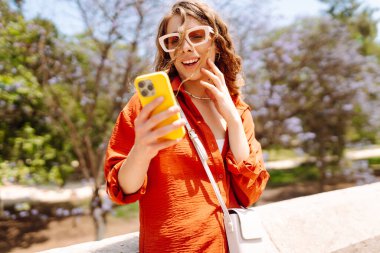 Şık güneş gözlüklü genç bir kadın güneşli bir günde canlı ağaçların yanında telefonunu kullanıyor. Seyahat, moda, iletişim, blog konsepti.