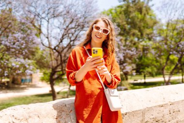Şık güneş gözlüklü genç bir kadın güneşli bir günde canlı ağaçların yanında telefonunu kullanıyor. Seyahat, moda, iletişim, blog konsepti.