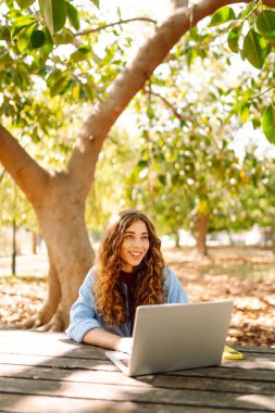 Serbest giyinmiş genç bir kadın güneşli bir günde park masasında oturmuş bir anlık üretkenliğin tadını çıkarırken dizüstü bilgisayara odaklandı. E-öğrenme konsepti. Uzak çalışma.