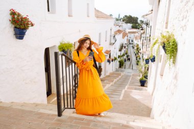 Turuncu elbiseli genç bir gezgin kadın güneşli bir köyde beyaz yıkanmış bir sokakta dolaşırken akıllı telefonunu kullanmaktan zevk alır. Seyahat, turizm, teknoloji, blog yazma.