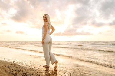 Gün batımında kıyıda sakin bir anın tadını çıkaran bir kadın. Hafif dalgalar ayaklarına vuruyor ve altın bir ufuk çiziyor. Yaz tatilindeki kadın turist.