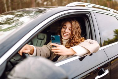 Kıvırcık saçlı genç bir kadın gülümseyerek araba sürmekten ve akıllı telefonunu kullanmaktan, güneşli bir günde neşe dolu bir anı yakalamaktan ve sıradan bir güven göstermekten hoşlanır. Boş zaman, seyahat, teknoloji. Araba paylaşımı.