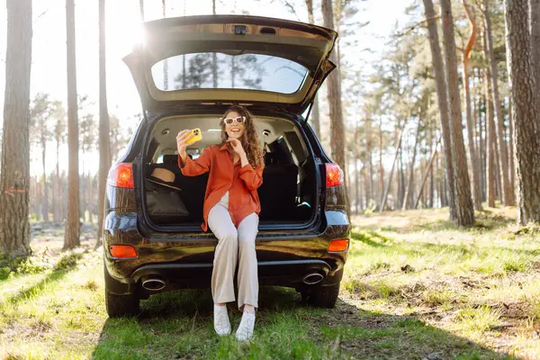 stock image A cheerful woman sits on the trunk of her car in a vibrant forest and capturing a selfie with her smartphone, radiating happiness. Travel, weekend, nature, relax and lifestyle concept.