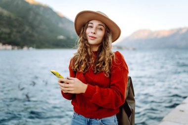 Genç bir kadın, deniz kenarındaki ahşap bir rıhtımda oturmuş güneşli bir günün tadını çıkarırken akıllı telefonunu güzel bir yerde kullanıyor. İnternetten alışveriş yapan, sosyal medyayı izleyen ya da mobil uygulamalarda sohbet eden kadınlar..