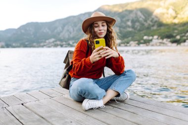 Genç bir kadın, deniz kenarındaki ahşap bir rıhtımda oturmuş güneşli bir günün tadını çıkarırken akıllı telefonunu güzel bir yerde kullanıyor. İnternetten alışveriş yapan, sosyal medyayı izleyen ya da mobil uygulamalarda sohbet eden kadınlar..