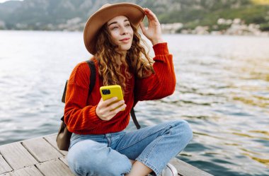 Genç bir kadın, deniz kenarındaki ahşap bir rıhtımda oturmuş güneşli bir günün tadını çıkarırken akıllı telefonunu güzel bir yerde kullanıyor. İnternetten alışveriş yapan, sosyal medyayı izleyen ya da mobil uygulamalarda sohbet eden kadınlar..