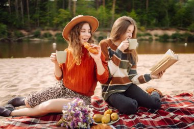 İki arkadaş güneşli bir öğleden sonra nehri kenarında piknik yaparken ellerinde çiçekler ve sıcak bir battaniyede atıştırmalıkları paylaşıyorlar. - Evet. Dişiler mini piknikte doğanın tadını çıkarırlar. Rahatlama kavramı, doğa..