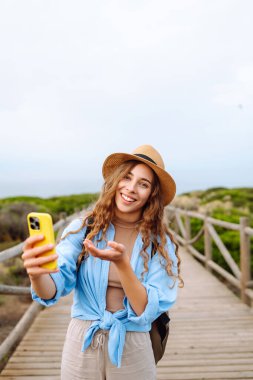 Kıvırcık saçlı genç kadın, açık bir gökyüzünün altında yeşil kıyı çalılıklarıyla çevrili bir selfie çekerken şık bir şapka takıyor ve el sallıyor. Seyahat, turizm, teknoloji, blog, iletişim kavramı.