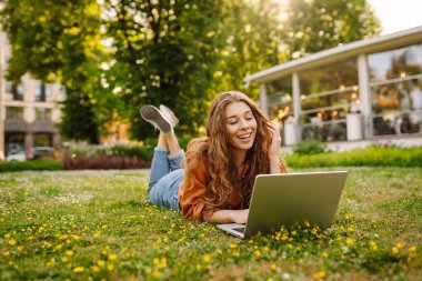 Genç bir kadın, doğa ve çiçeklerle çevrili çimlerin üzerinde rahatça otururken bilgisayarıyla meşgul olarak parkta güneşli bir öğleden sonranın tadını çıkarıyor. Serbest Çalışan, Çevrimiçi Eğitim.
