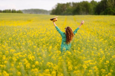 A woman in a green floral dress enjoys a sunny day in a vibrant yellow flower field near a forest during springtime. Nature, fashion, summer lifestyle. clipart