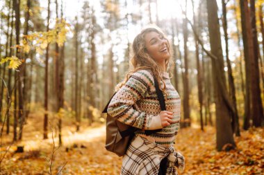Genç bir kadın, uzun ağaçlar ve düşen sonbahar yapraklarıyla çevrili, kolları açık, mutluluk saçan güneşli bir ormanda duruyor. Sonbahar manzarası
