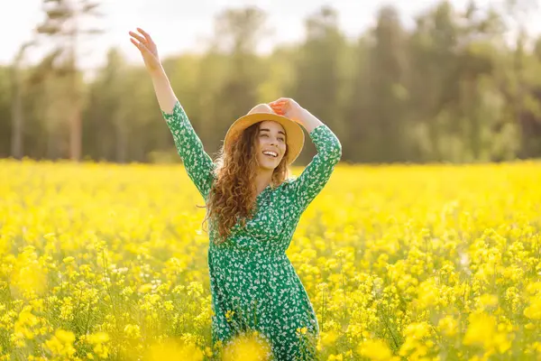 Yeşil çiçekli bir elbise giyen bir kadın, bahar zamanı bir ormanın yakınındaki canlı sarı bir çiçek tarlasında güneşli bir günün tadını çıkarır. Doğa, moda, yaz yaşam tarzı.