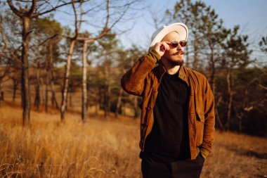 Güneş gözlüğü takan ve büyük şapkalı bir adam sonbahar boyunca ağaçlarla çevrili çimenli bir alanda güneşli bir günün tadını çıkarıyor. Sonbahar manzarası.