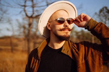 Güneş gözlüğü takan ve büyük şapkalı bir adam sonbahar boyunca ağaçlarla çevrili çimenli bir alanda güneşli bir günün tadını çıkarıyor. Sonbahar manzarası.