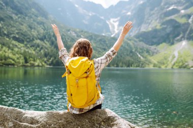 Sırt çantalı genç bir kadın, berrak bir göl kenarındaki kayalık arazide poz verirken gülümsüyor. Parlak bir gökyüzünün altındaki görkemli dağlar ve yemyeşil ağaçlarla çerçevelenmiş. Etkin yaşam biçimi.