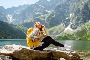 Sırt çantalı genç bir kadın, berrak bir göl kenarındaki kayalık arazide poz verirken gülümsüyor. Parlak bir gökyüzünün altındaki görkemli dağlar ve yemyeşil ağaçlarla çerçevelenmiş. Etkin yaşam biçimi.