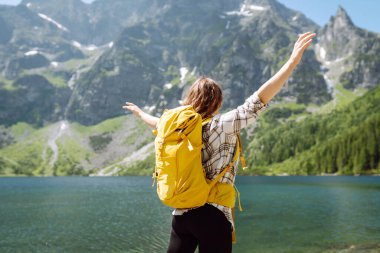 Sırt çantalı genç bir kadın, berrak bir göl kenarındaki kayalık arazide poz verirken gülümsüyor. Parlak bir gökyüzünün altındaki görkemli dağlar ve yemyeşil ağaçlarla çerçevelenmiş. Etkin yaşam biçimi.