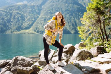 Sırt çantalı genç bir kadın, berrak bir göl kenarındaki kayalık arazide poz verirken gülümsüyor. Parlak bir gökyüzünün altındaki görkemli dağlar ve yemyeşil ağaçlarla çerçevelenmiş. Etkin yaşam biçimi.