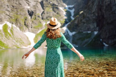 Bir kadın, yeşil bir elbise ve geniş kenarlı bir şapka giymiş, arka planda çarpıcı dağlar bulunan sakin bir dağ gölünde yalınayak dolaşır. Moda, güzellik, yaşam tarzı, seyahat.