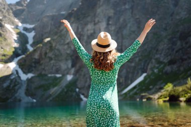 Bir kadın, yeşil bir elbise ve geniş kenarlı bir şapka giymiş, arka planda çarpıcı dağlar bulunan sakin bir dağ gölünde yalınayak dolaşır. Moda, güzellik, yaşam tarzı, seyahat.