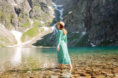 Bir kadın, yeşil bir elbise ve geniş kenarlı bir şapka giymiş, arka planda çarpıcı dağlar bulunan sakin bir dağ gölünde yalınayak dolaşır. Moda, güzellik, yaşam tarzı, seyahat.