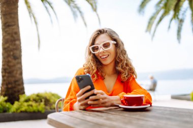 Güneş gözlüklü bir kadın, palmiye ağaçlarıyla çevrili güneşli bir kafede akıllı telefonunu kullanırken kahveden zevk alır. İnsanlar, moda, yaşam tarzı, seyahat ve tatil kavramı.