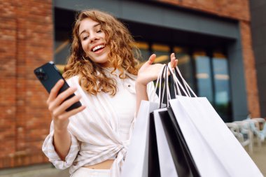 Genç bir kadın, canlı bir kentsel bölgede güneşli bir günde bir mağazanın önünde telefonunu kontrol ederken alışveriş yapmaktan zevk alıyor. Tüketim, satış, kara cuma. İnternetten alışveriş.