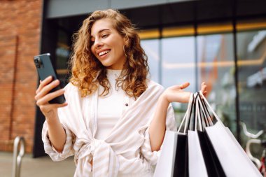 Genç bir kadın, canlı bir kentsel bölgede güneşli bir günde bir mağazanın önünde telefonunu kontrol ederken alışveriş yapmaktan zevk alıyor. Tüketim, satış, kara cuma. İnternetten alışveriş.