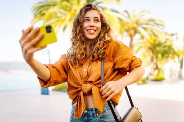Neşeli genç bir kadın, suyun kenarında güneşli bir günün tadını çıkarıyor. Parlak sarı bir kamerayla selfie çekiyor ve her tarafı gür palmiye ağaçlarıyla çevrili. Seyahat günlüğü