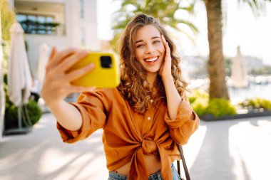 Neşeli genç bir kadın, suyun kenarında güneşli bir günün tadını çıkarıyor. Parlak sarı bir kamerayla selfie çekiyor ve her tarafı gür palmiye ağaçlarıyla çevrili. Seyahat günlüğü