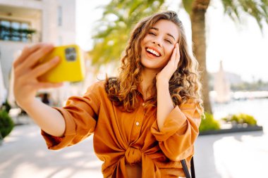 Neşeli genç bir kadın, suyun kenarında güneşli bir günün tadını çıkarıyor. Parlak sarı bir kamerayla selfie çekiyor ve her tarafı gür palmiye ağaçlarıyla çevrili. Seyahat günlüğü