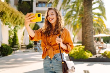 Neşeli genç bir kadın, suyun kenarında güneşli bir günün tadını çıkarıyor. Parlak sarı bir kamerayla selfie çekiyor ve her tarafı gür palmiye ağaçlarıyla çevrili. Seyahat günlüğü