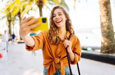 Neşeli genç bir kadın, suyun kenarında güneşli bir günün tadını çıkarıyor. Parlak sarı bir kamerayla selfie çekiyor ve her tarafı gür palmiye ağaçlarıyla çevrili. Seyahat günlüğü