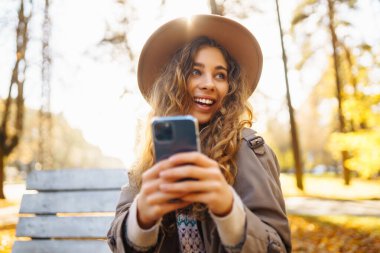Parkta akıllı telefonuyla mesajlaşırken güneşli bir sonbahar gününün tadını çıkaran genç bir kadın. Tatil kavramı, blog yazma, teknoloji, hafta sonu.