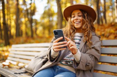 Parkta akıllı telefonuyla mesajlaşırken güneşli bir sonbahar gününün tadını çıkaran genç bir kadın. Tatil kavramı, blog yazma, teknoloji, hafta sonu.