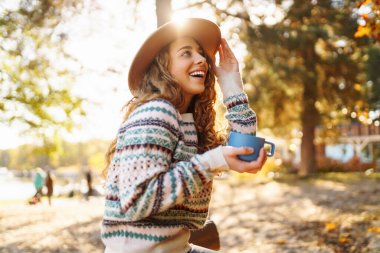 Genç bir kadın hareketli ağaçlarla çevrili güneşli bir sonbahar öğleden sonrasında dingin göl kıyısında kahvesini içiyor. Nehir kıyısındaki utumn manzarası. Seyahat konsepti, rahatla..