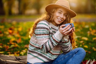 Sonbahar boyunca parkta, renkli yapraklarla çevrili sıcak bir içeceğin keyfini çıkaran genç bir bayanla sıcak öğleden sonra pikniği. Dinlenme, rahatlama kavramı. Yaşam biçimi.