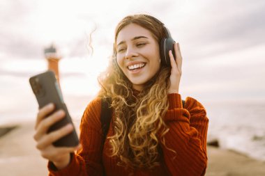 Genç bir kadın, altın saat boyunca deniz kenarındaki akıllı telefonuyla müzik dinlemekten zevk alıyor, kulaklık takıyor ve neşeyle gülümsüyor. Seyahat, turizm, teknoloji, blog konsepti.