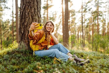 Mutlu genç bir kadın, güneşli bir günde ormanlık alanda bir ağacın yanında otururken selfie çekiyor. Seyahat blogu. Macera, tatil.