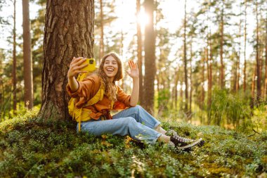 Mutlu genç bir kadın, güneşli bir günde ormanlık alanda bir ağacın yanında otururken selfie çekiyor. Seyahat blogu. Macera, tatil.