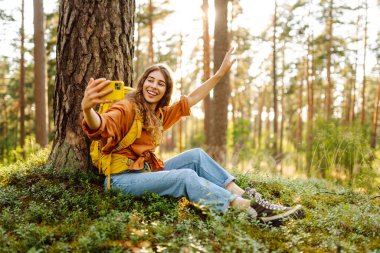 Mutlu genç bir kadın, güneşli bir günde ormanlık alanda bir ağacın yanında otururken selfie çekiyor. Seyahat blogu. Macera, tatil.