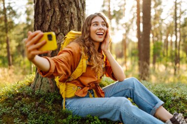 Mutlu genç bir kadın, güneşli bir günde ormanlık alanda bir ağacın yanında otururken selfie çekiyor. Seyahat blogu. Macera, tatil.